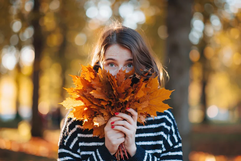 Young woman holding bouquet of orange maple leaves in front of her face in autumn park