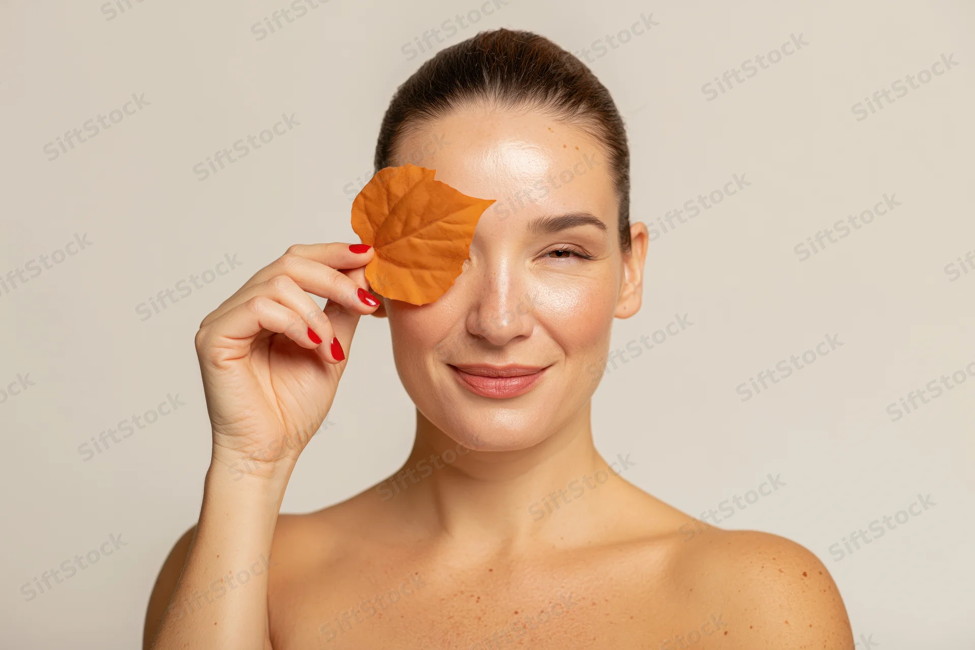 Woman holding orange autumn leaf over one eye in studio portrait