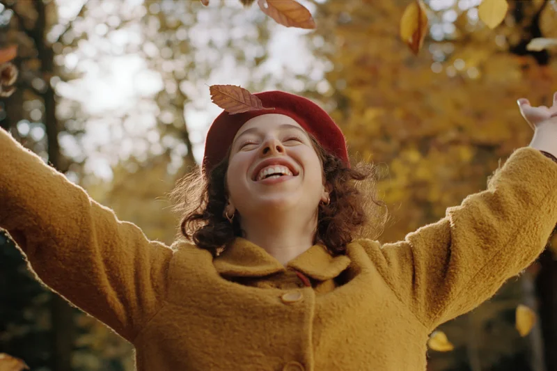 Young woman laughing with arms raised amidst falling autumn leaves in a sunlit forest