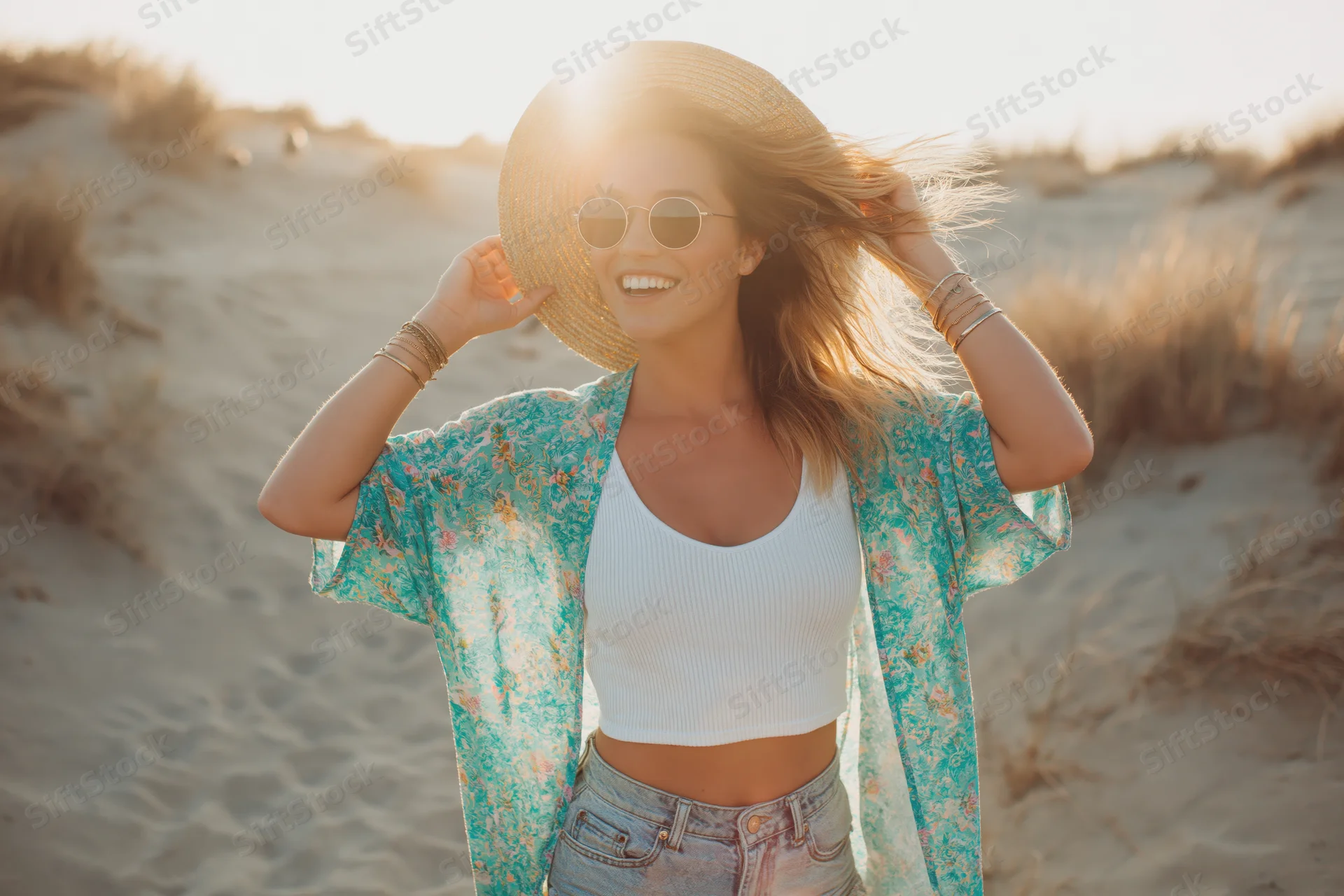 Woman laughing on beach holding straw hat to her head at sunset