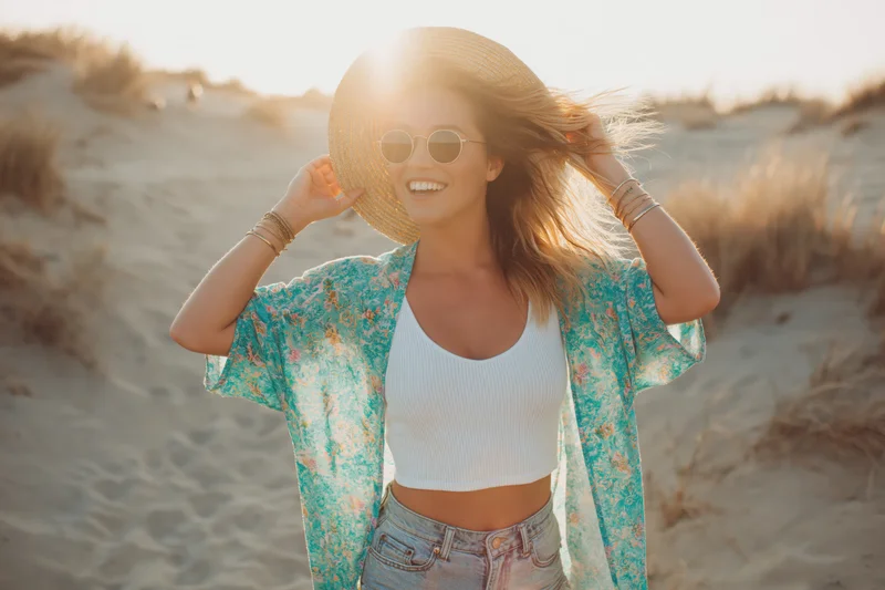 Woman laughing on beach holding straw hat to her head at sunset