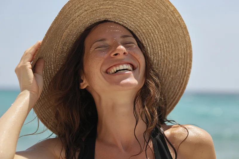 Woman laughing with eyes closed on beach wearing straw hat