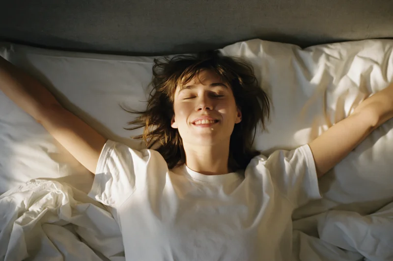 Young woman lying on back in bed stretching arms wide with eyes closed and smiling