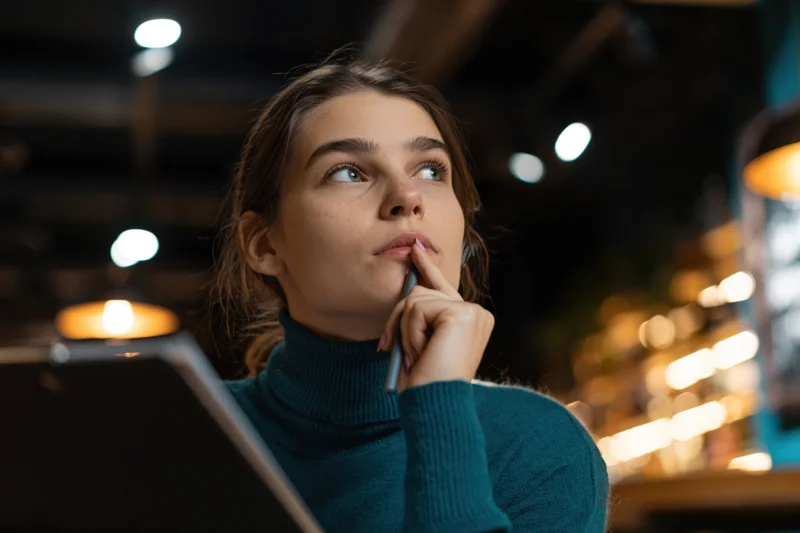 Woman thinking with pen to lips in dim cafe lounge with laptop open before her