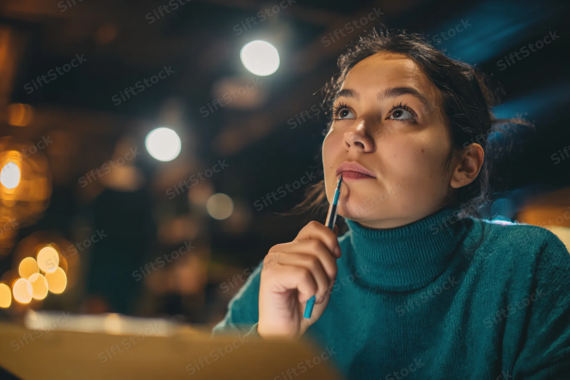 Young woman holding pencil to chin while thinking in dim cafe lounge at night