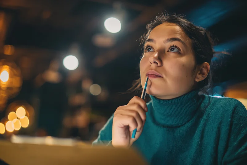 Young woman holding pencil to chin while thinking in dim cafe lounge at night