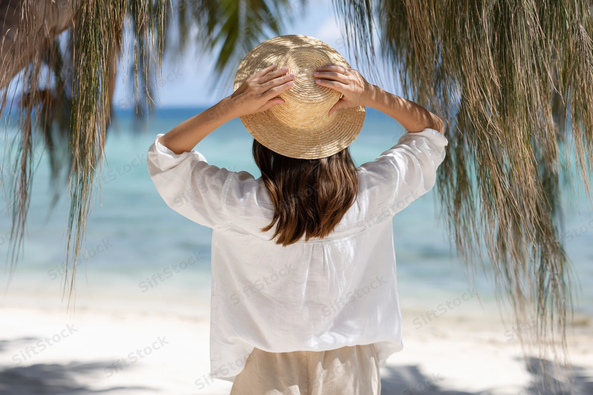 Woman from behind holding straw hat on tropical beach under palm fronds
