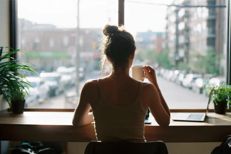 Woman sitting at wooden cafe counter holding mug with laptop and plants, looking out window at city street