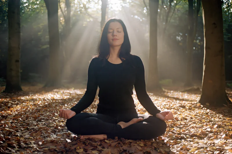 Woman meditating in lotus pose on forest floor covered in autumn leaves with sunlight filtering through trees