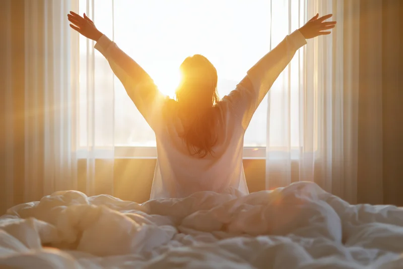 Woman stretching arms overhead in bed by sunrise window