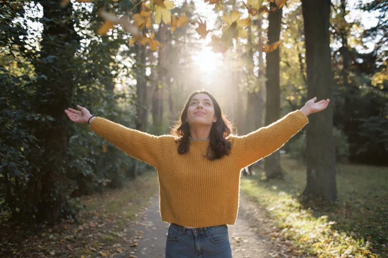 Woman with arms outstretched tossing autumn leaves in a sunlit forest path