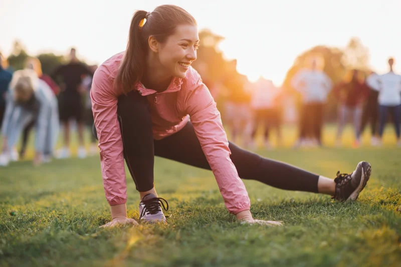 Woman stretching her leg on grass during sunset in a park with others exercising in the background