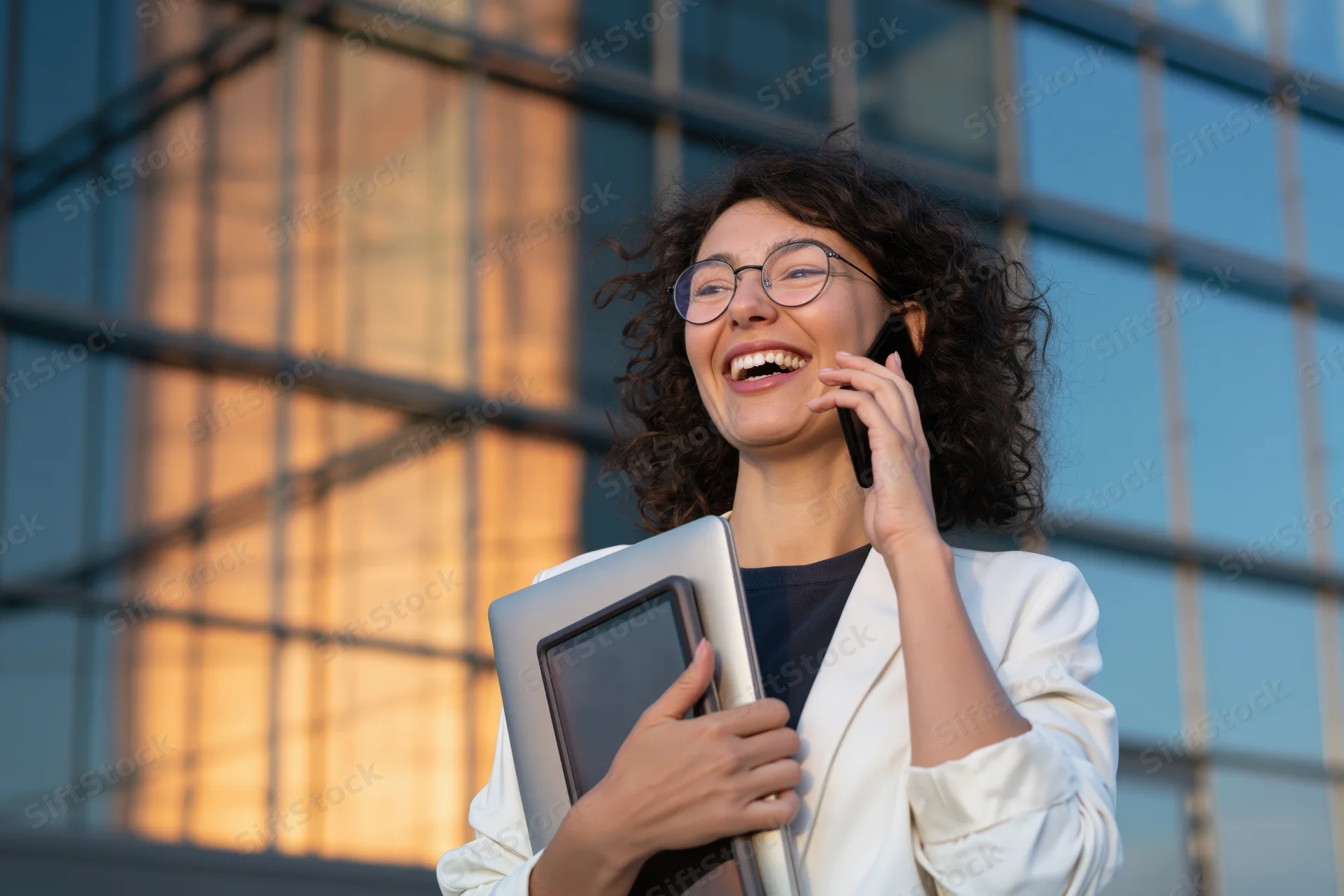 Woman laughing while talking on phone and holding laptop outside modern office building at sunset