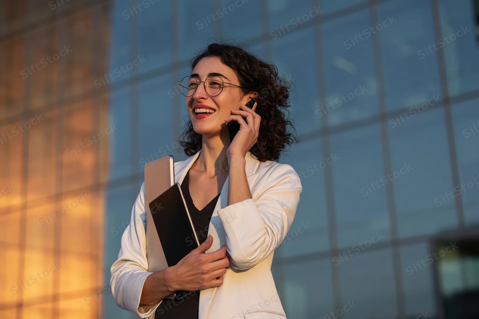Woman laughing while talking on smartphone outside modern glass building at sunset