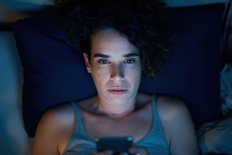Woman lying in bed at night holding phone, illuminated by screen light