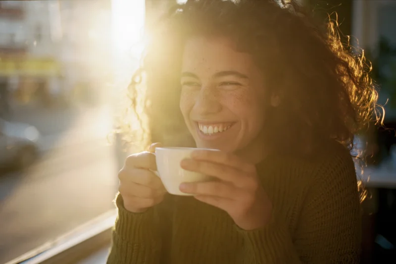 Young woman laughing while holding a white coffee cup by a sunlit window