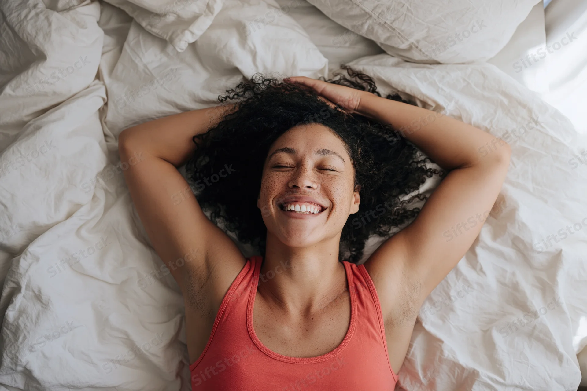 Woman smiling with eyes closed lying on her back in bed with arms behind head