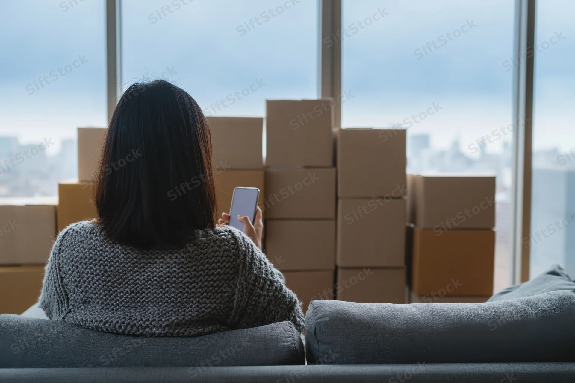 Woman sitting on couch using smartphone with moving boxes in front of city view window