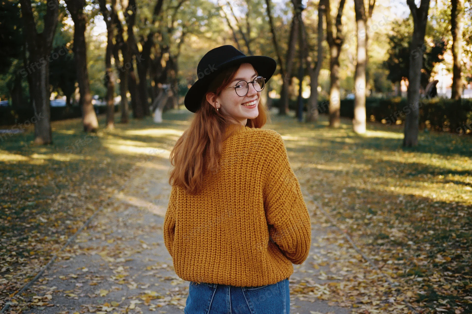 Woman in mustard sweater and black hat smiling over shoulder in autumn park