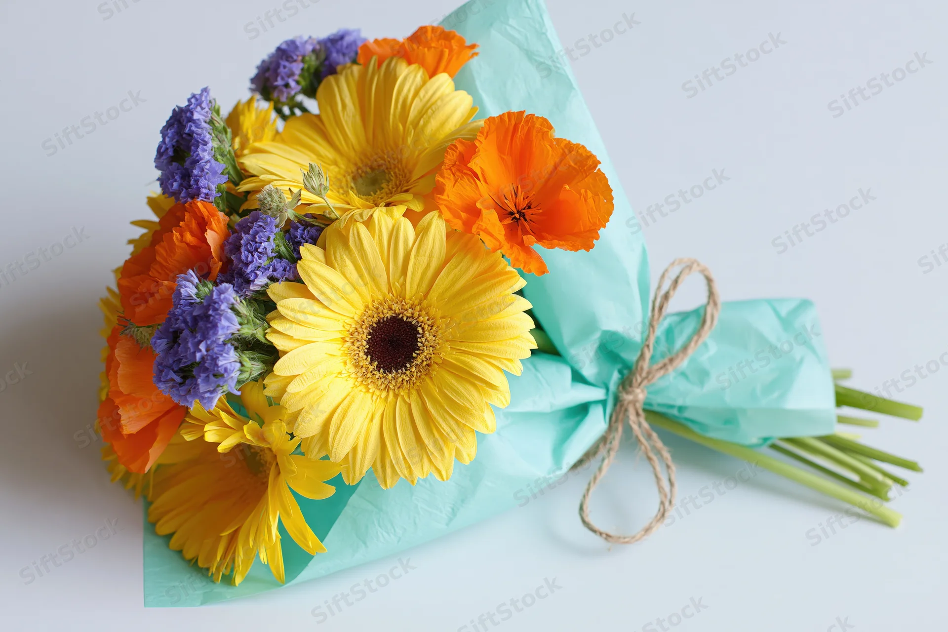 Hand-tied bouquet of yellow gerbera daisies, orange poppies, and purple statice wrapped in teal paper with twine on white surface
