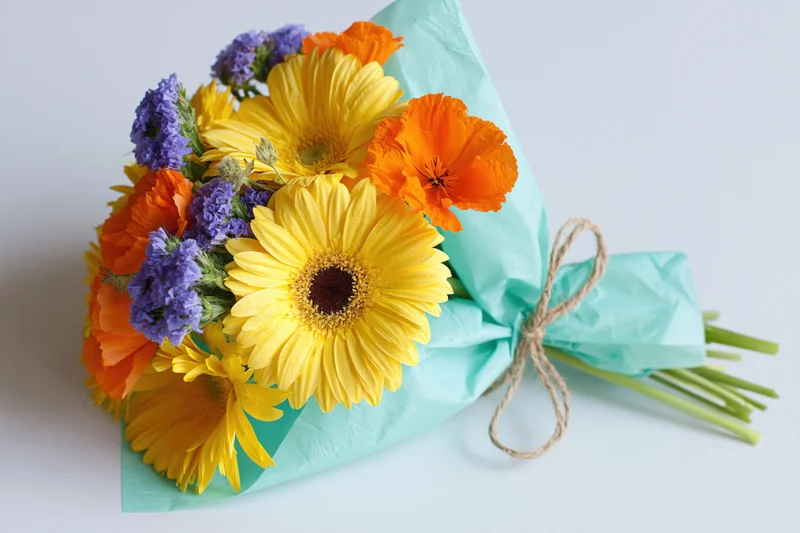 Hand-tied bouquet of yellow gerbera daisies, orange poppies, and purple statice wrapped in teal paper with twine on white surface