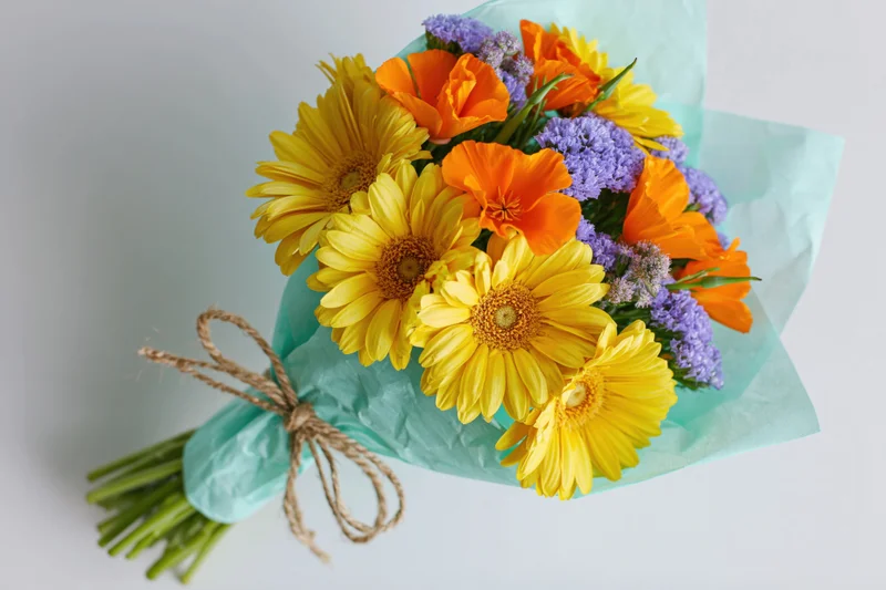 Hand-tied bouquet of yellow gerbera daisies, orange poppies, and purple lavender wrapped in teal paper with twine on white background
