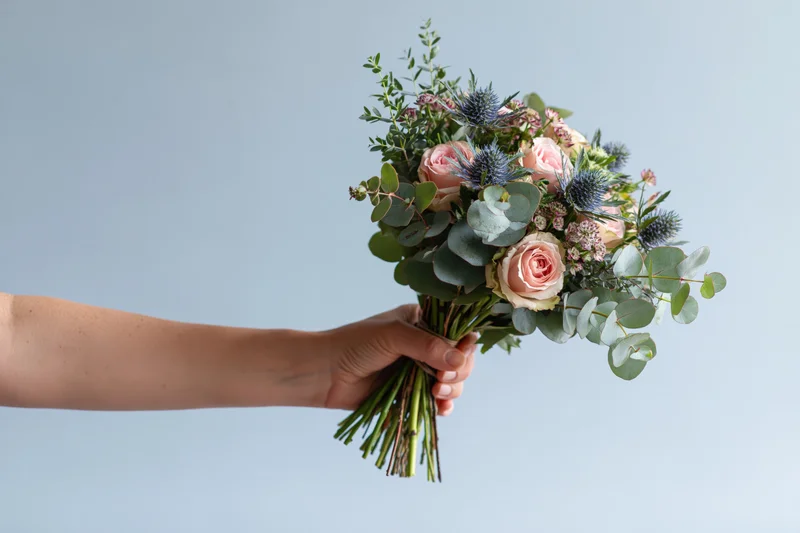Hand holding a bouquet of pink roses, blue thistle and eucalyptus against a light background