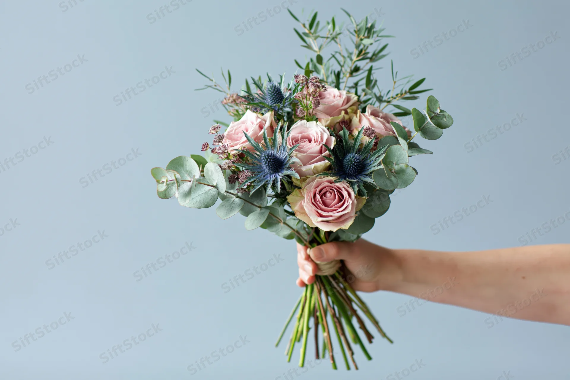 Hand holding a bouquet of pink roses, blue thistle and eucalyptus against a light background