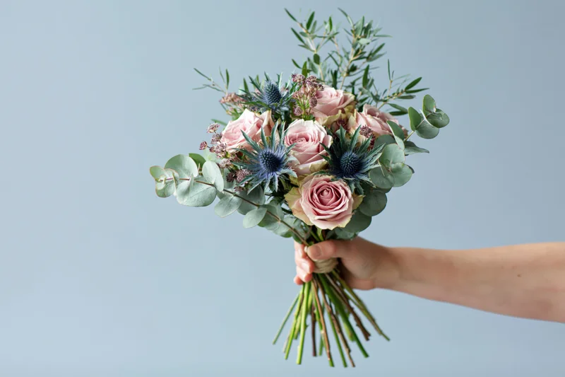 Hand holding a bouquet of pink roses, blue thistle and eucalyptus against a light background