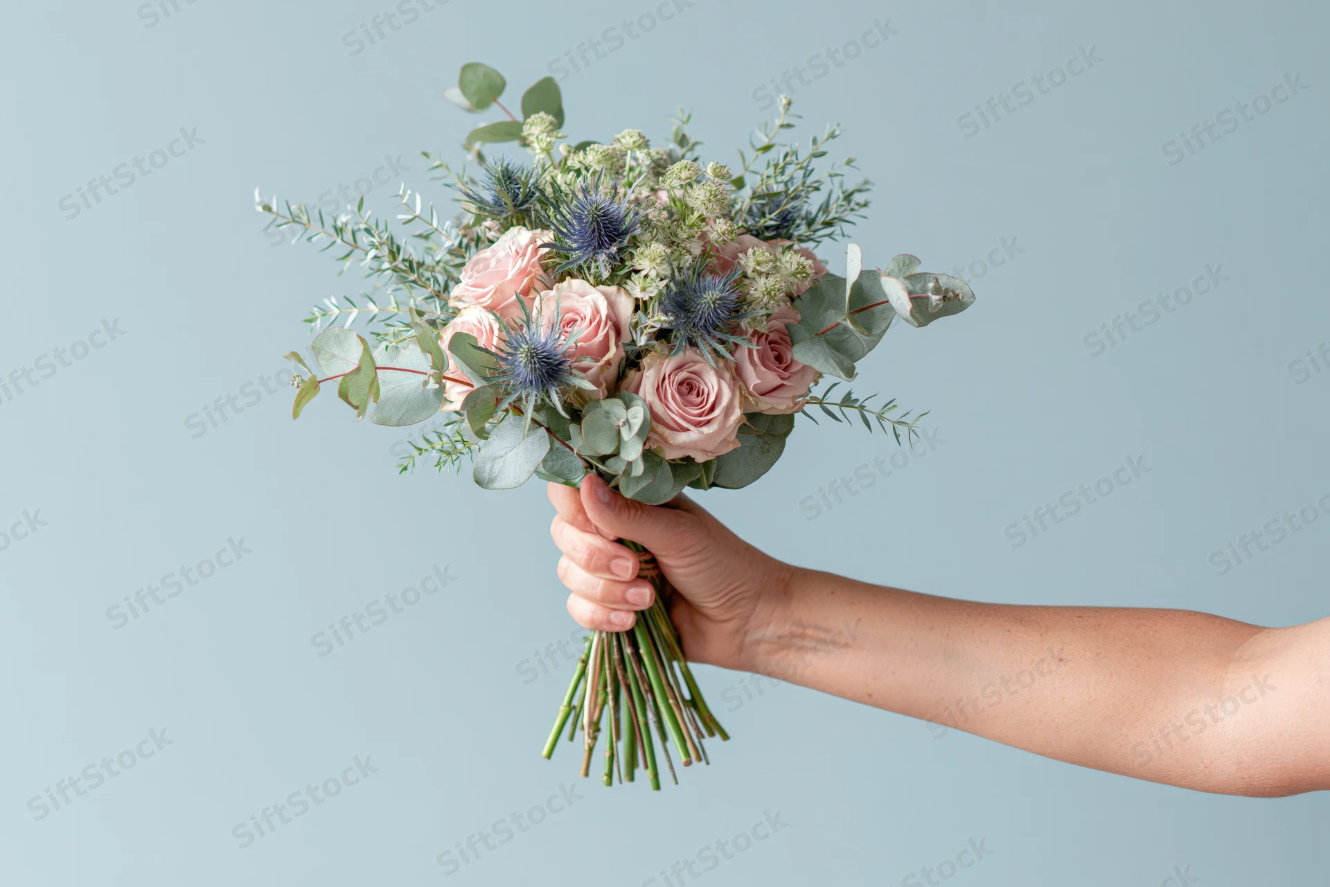 Hand holding a bouquet of pink roses, blue thistle and eucalyptus against a light blue background