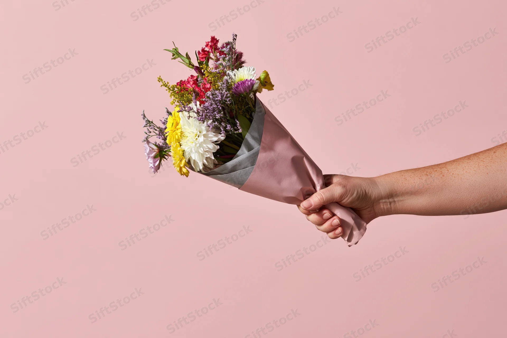 Hand holding a mixed flower bouquet wrapped in pink paper against a pink background