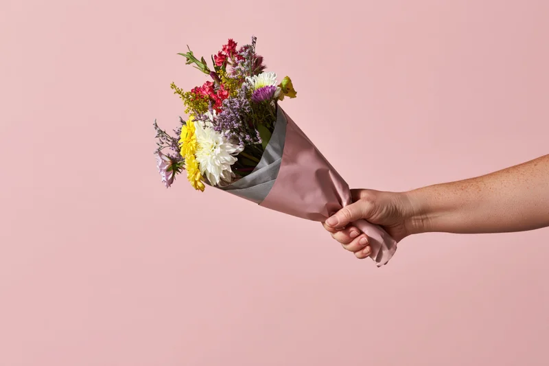 Hand holding a mixed flower bouquet wrapped in pink paper against a pink background