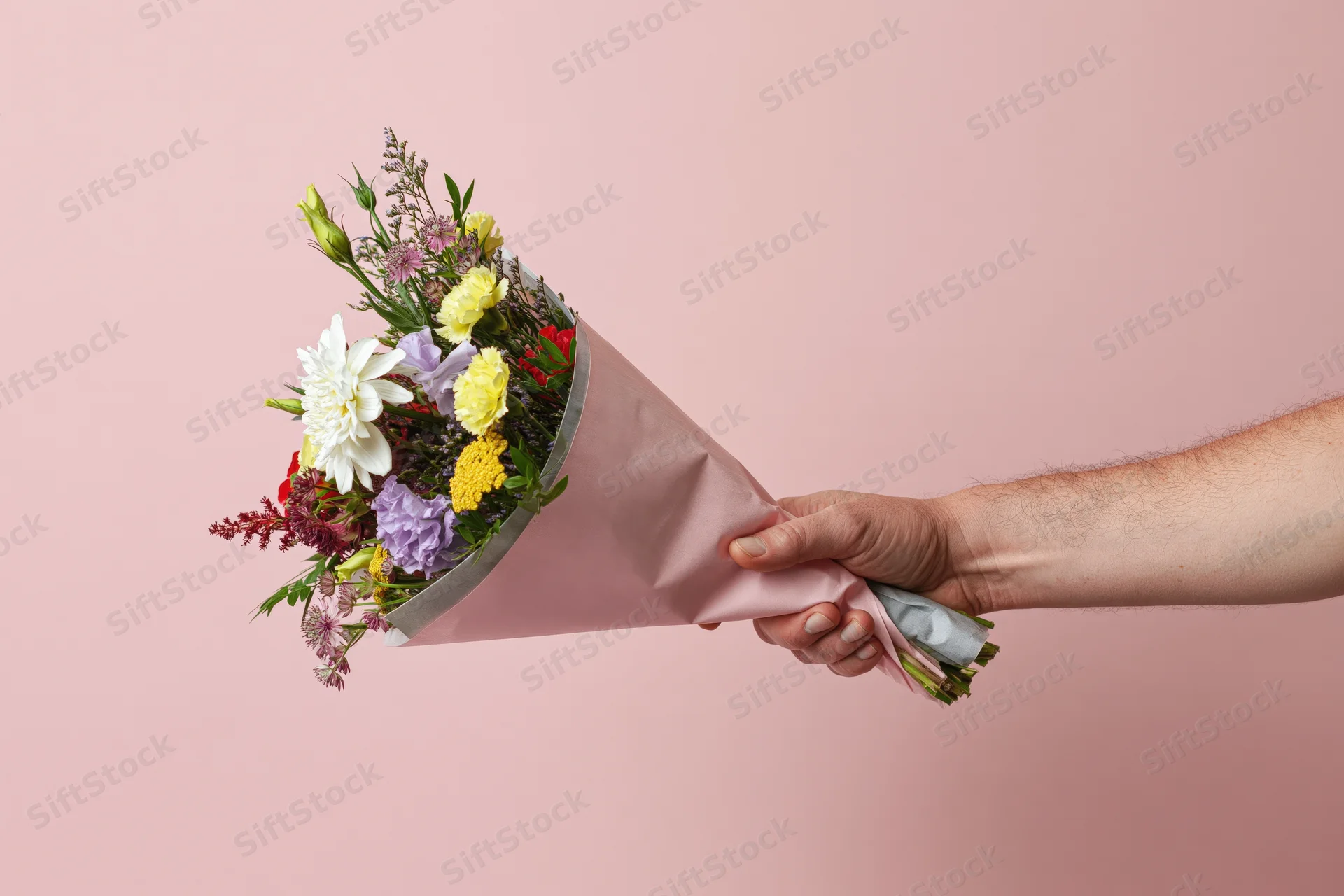 Hand holding a mixed flower bouquet wrapped in pink paper against a pink background