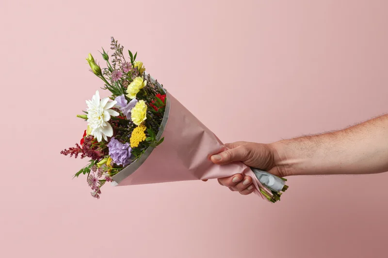 Hand holding a mixed flower bouquet wrapped in pink paper against a pink background