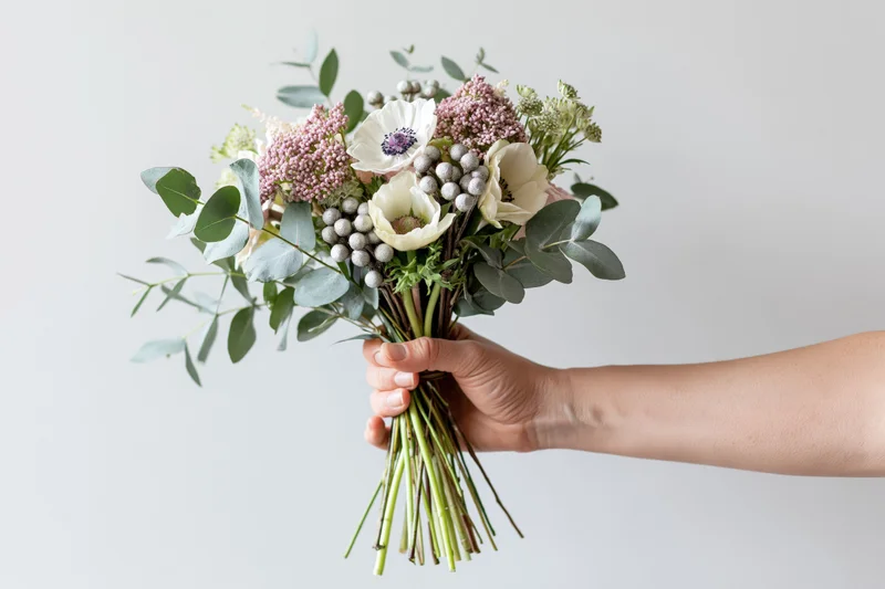 Hand holding a mixed floral bouquet with white anemones and eucalyptus against a light background