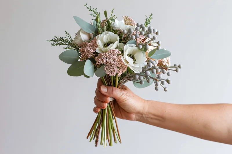 Hand holding a mixed bouquet of white anemones, pink astilbe, eucalyptus and greenery against a plain background