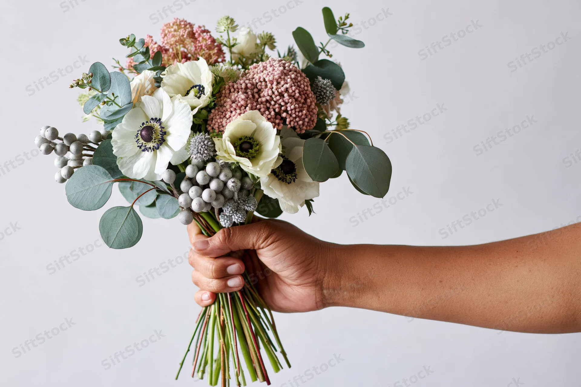 Hand holding a mixed floral bouquet with white anemones and greenery against a plain background