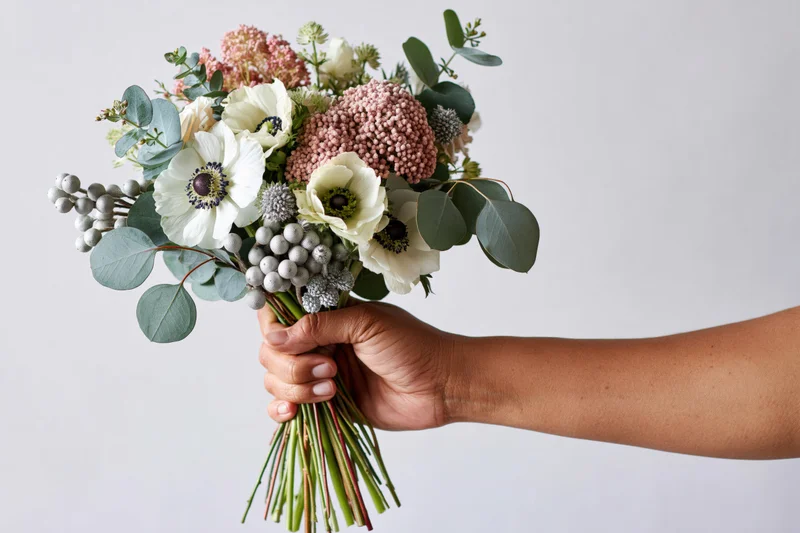 Hand holding a mixed floral bouquet with white anemones and greenery against a plain background