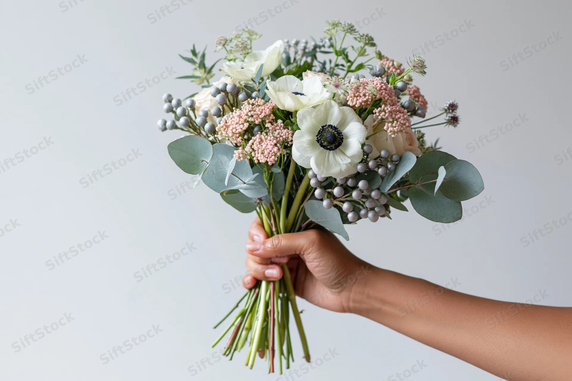 Hand holding a mixed bouquet of white anemones, pink ranunculus, and eucalyptus against a plain background