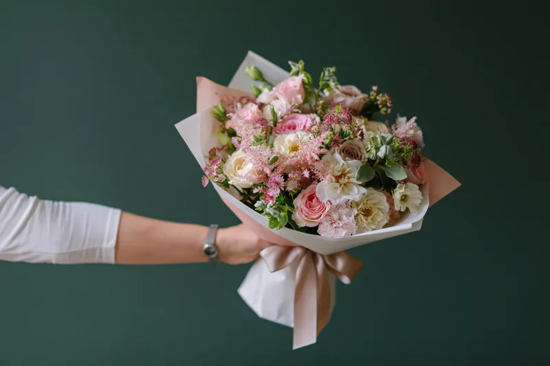 Woman's hand holding a wrapped bouquet of pink and cream flowers against a dark green background