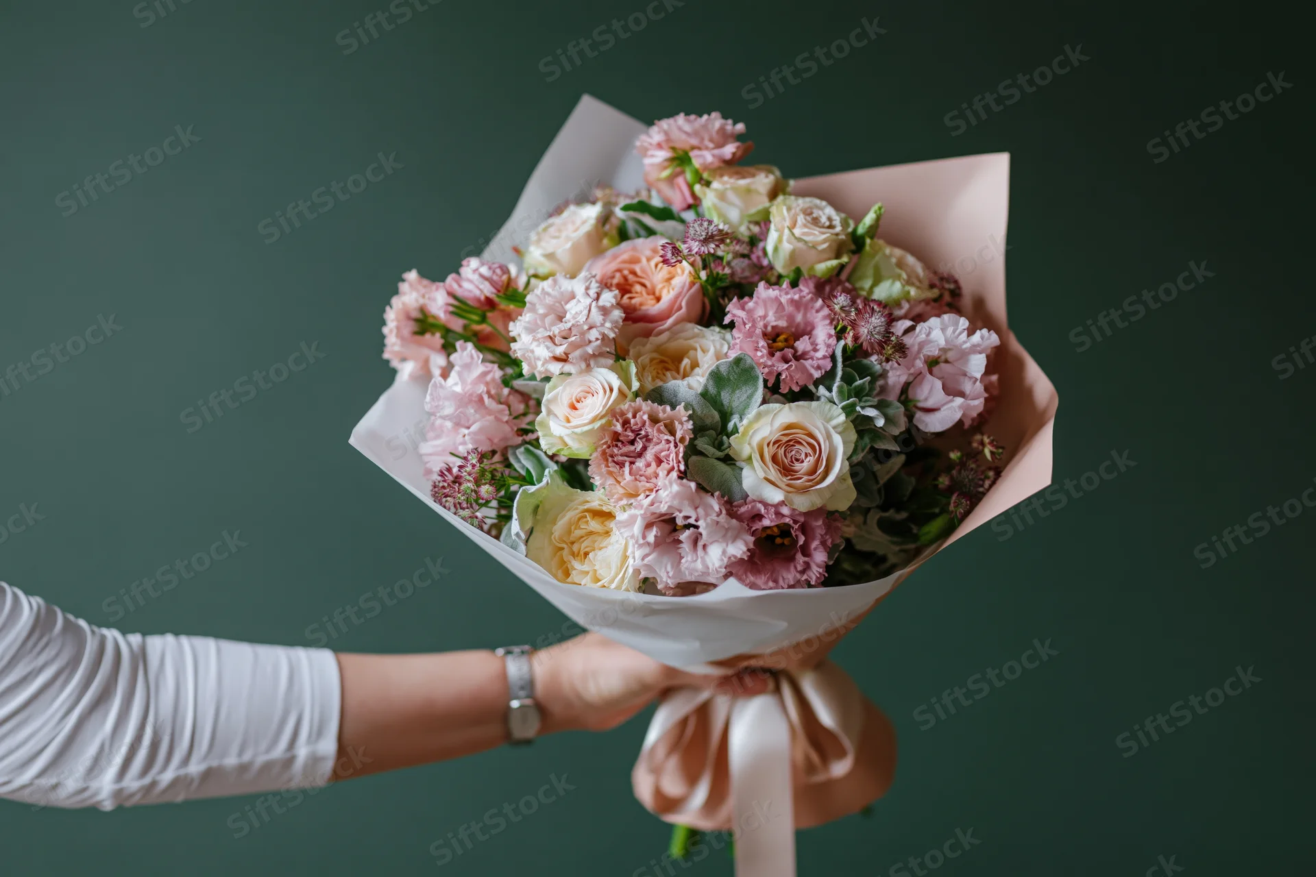 Woman's hand holding a wrapped bouquet of pink and cream roses and carnations against a dark green background