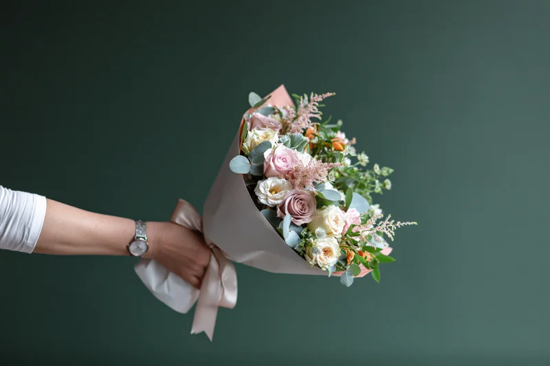 Woman's hand holding a wrapped bouquet of pink and white roses with greenery against a dark green background