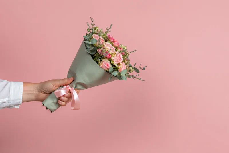 Hand in white shirt sleeve holding a pink rose bouquet wrapped in green paper with ribbon against a pink background