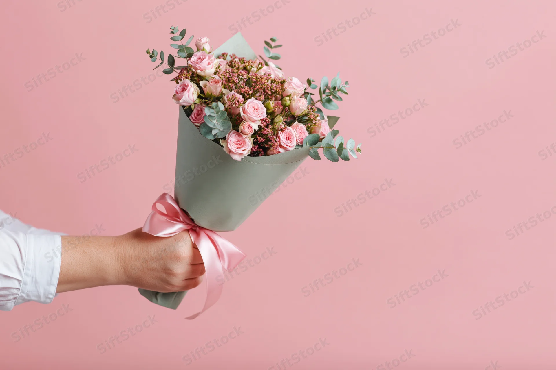 Hand holding a wrapped bouquet of pink roses and greenery against a pink background