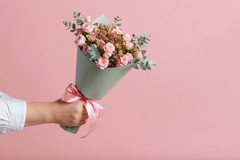 Hand holding a wrapped bouquet of pink roses and greenery against a pink background