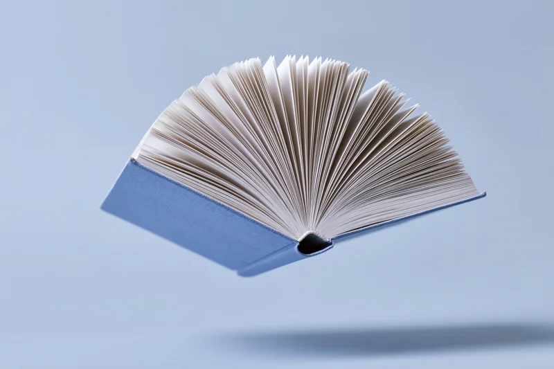 Open hardcover book floating mid-air with pages fanned out against a light blue background