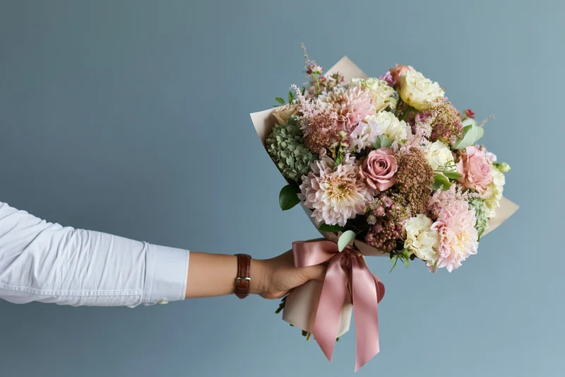 Woman's arm holding a wrapped bouquet of pink and white flowers against a blue background