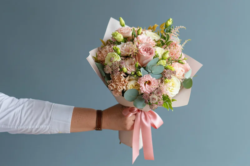 Hand holding a wrapped bouquet of pink and white flowers against a blue background