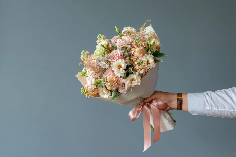 Hand holding a wrapped bouquet of pink and white flowers with greenery and ribbon against a gray background