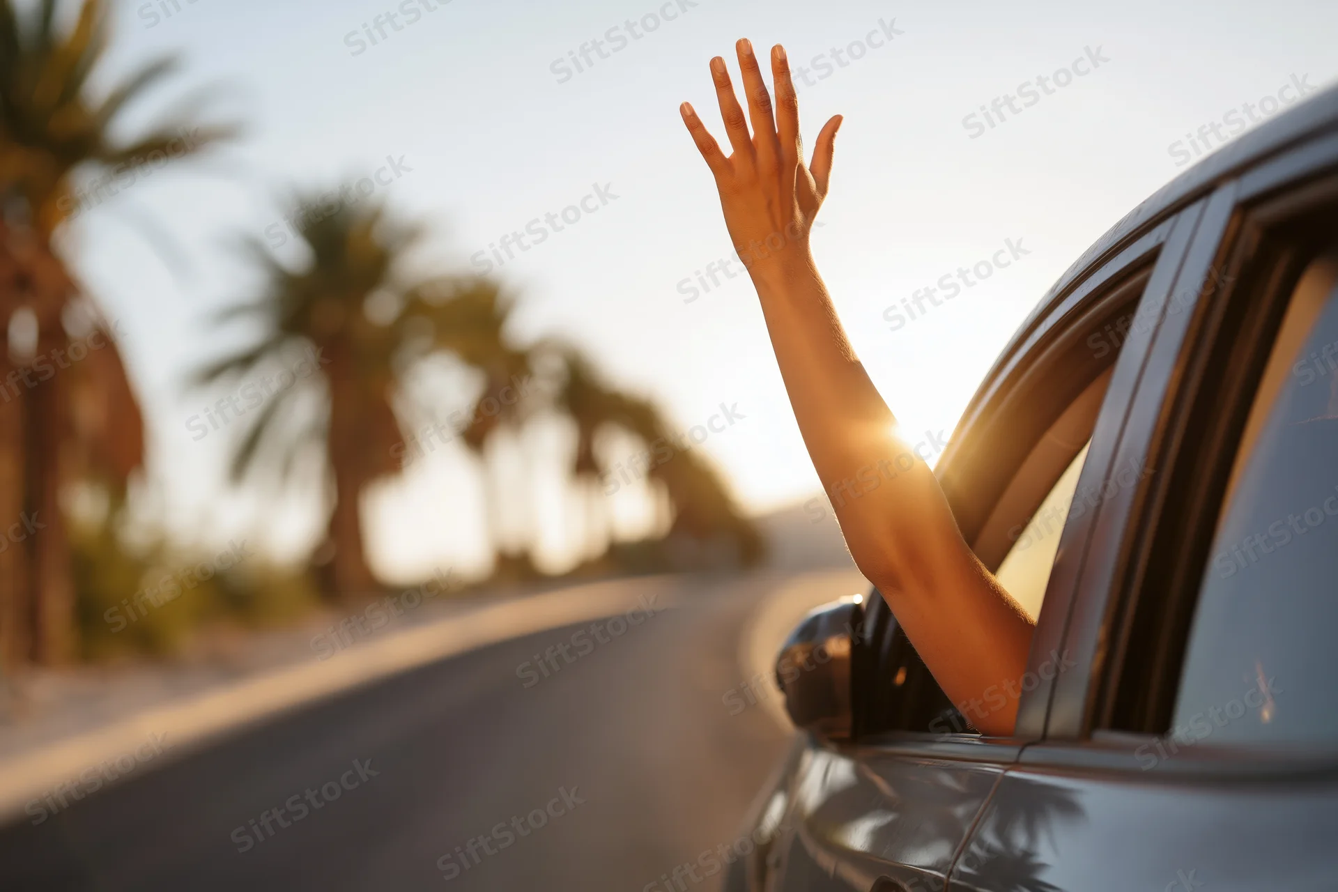 Woman’s arm reaching out of car window with hand raised on coastal road at sunset